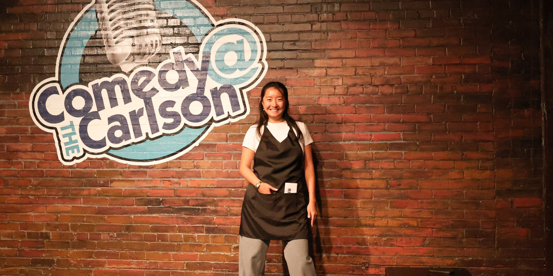 Person wearing an apron standing in front of a 'Comedy at the Carlson' sign on a brick wall.